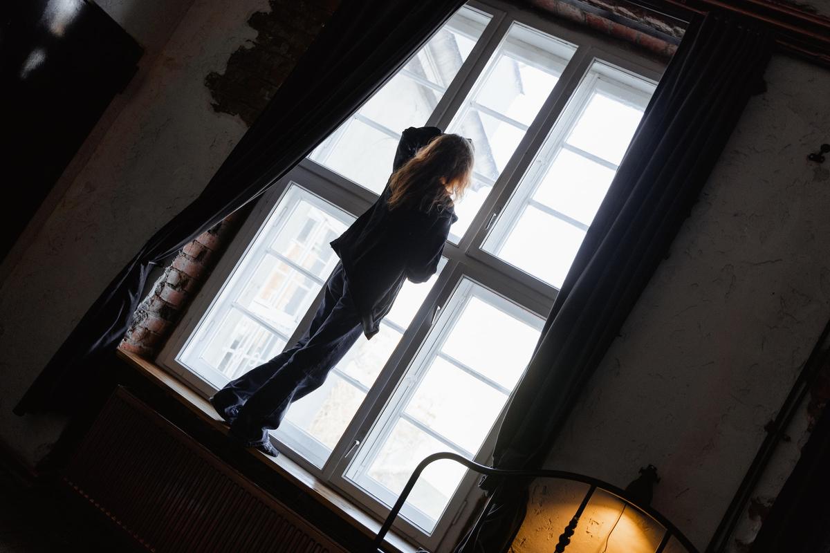 A high-angle, slightly tilted shot of a person with long blonde hair standing on a wide wooden windowsill, silhouetted against a large, multi-paned window.