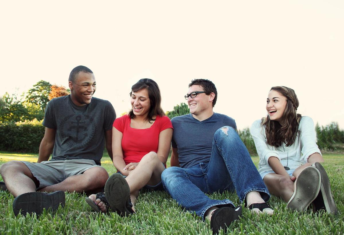 A diverse group of young adults sitting on grass outdoors, laughing and talking together, symbolizing connection, peer support, and building community during rehab and recovery.
