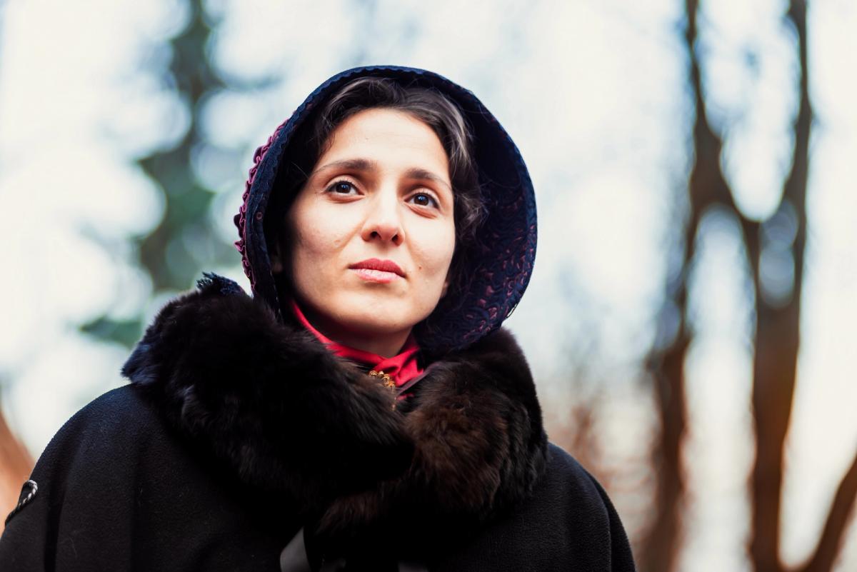 A woman in 19th-century attire, including a fur-trimmed cloak and a patterned bonnet, looking upward with a thoughtful expression.