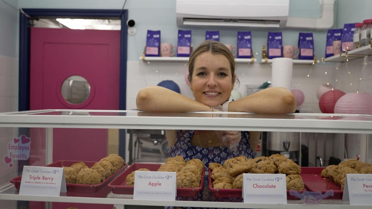 Janie Deegan leans on her elbows behind a bakery display case. The glass case features trays of "Pie Crust Cookies" in flavors like Triple Berry, Apple, and Chocolate.