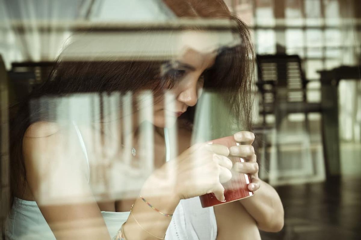 Thoughtful woman holding a mug of tea behind a window with reflections, sitting indoors and looking down in a quiet, reflective moment.