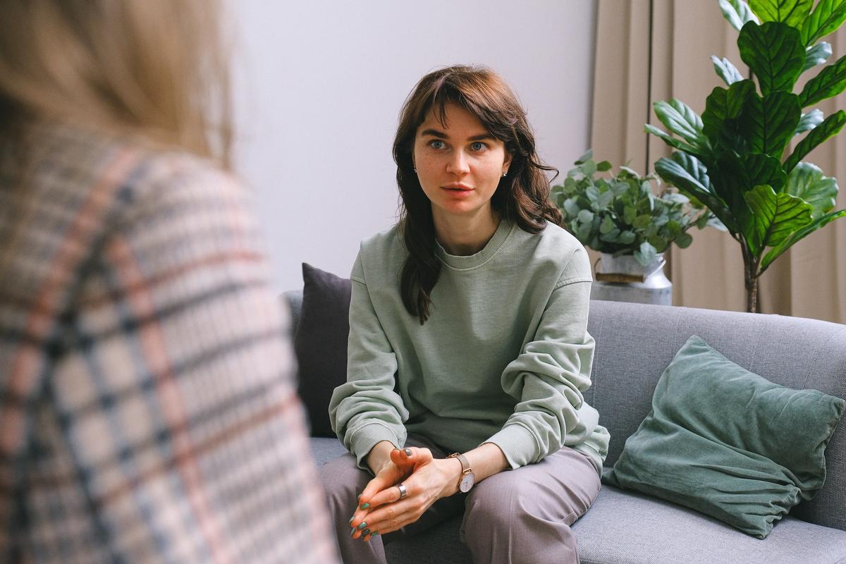 A woman sitting on a grey sofa looks intently at someone off-camera during a conversation.