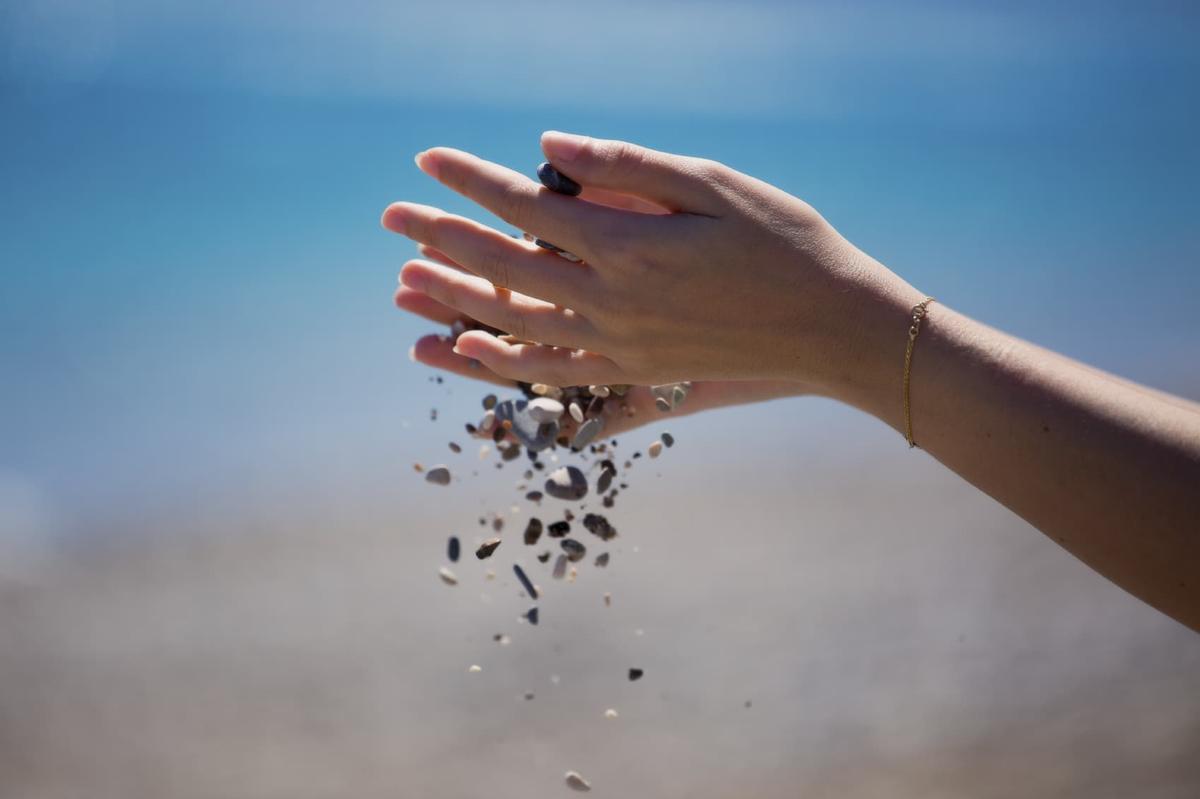 A person’s hands cupped together as small pebbles and sand fall through their fingers at a beach.