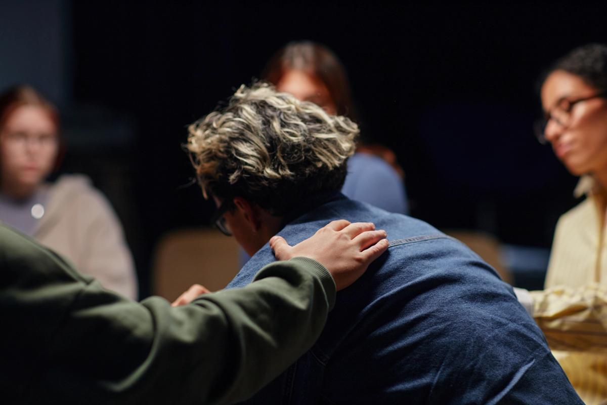 Person sitting in a support group with head lowered while another member places a reassuring hand on their shoulder, representing support for someone struggling with polysubstance use.