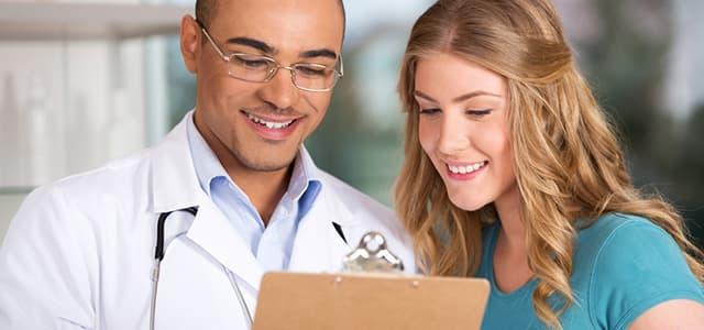 A close-up image of a smiling male doctor wearing glasses and a white lab coat with a stethoscope around his neck, standing next to a young female patient.