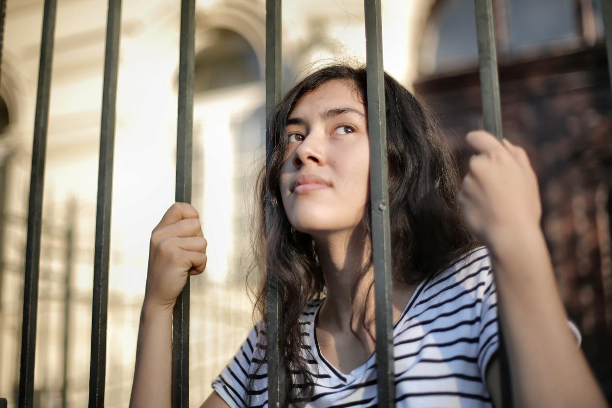 A young woman holding onto metal bars, looking upward with a thoughtful expression, suggesting confinement, reflection, or longing for freedom.