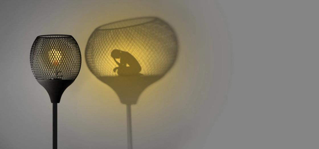 A lamp casts a large shadow on a wall, within which the silhouette of a person sitting in a crouched, pensive position appears.