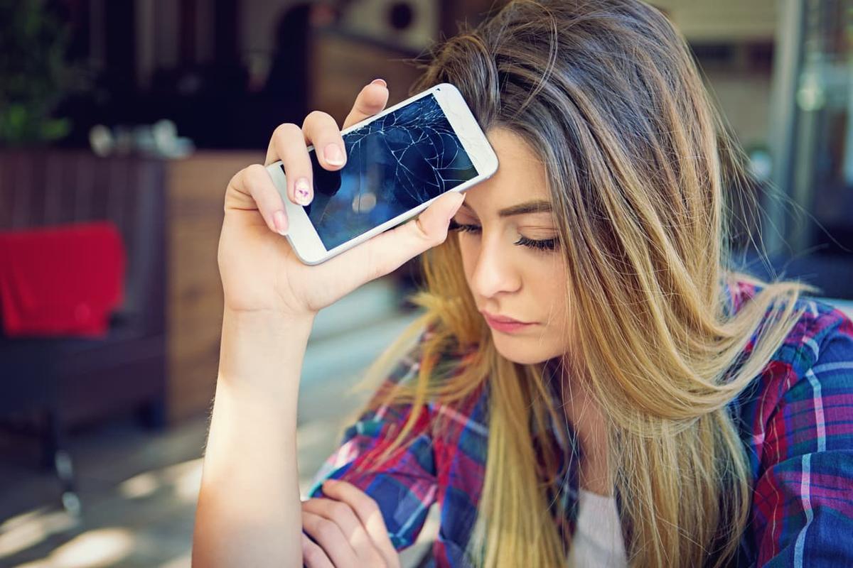 A young woman with long blonde hair looking down with a sad, stressed expression while holding a white smartphone with a severely cracked screen against her forehead.