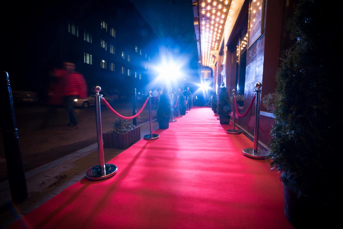 Red carpet entrance outside a building at night, lined with velvet ropes and bright spotlights, creating a glamorous event walkway.