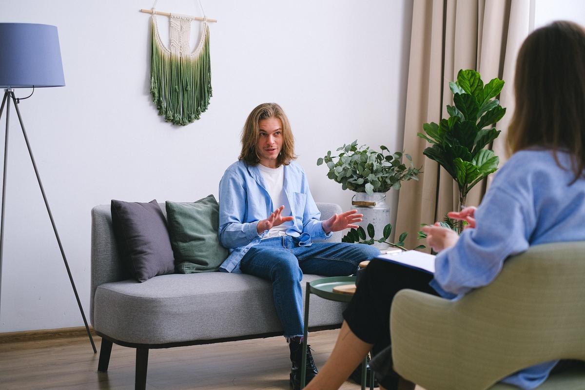 An eye-level shot of a young man with shoulder-length blonde hair sitting on a grey sofa, gesturing with his hands as he speaks during a therapy session.