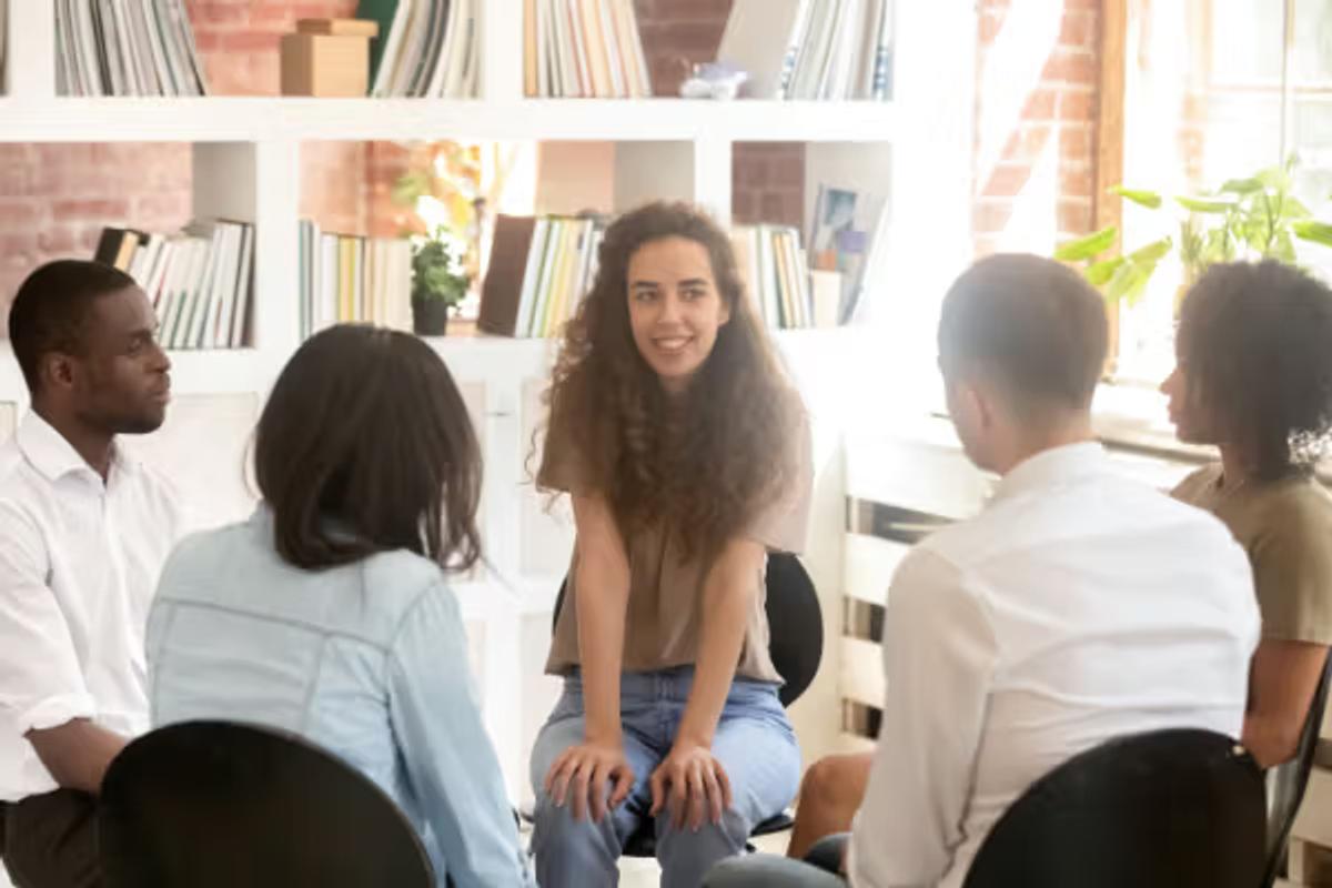A young woman with long, curly brown hair is sitting and speaking to a diverse group of four other adults seated in a circle around her.