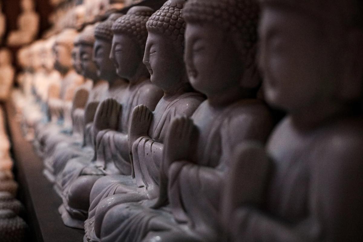 A row of identical stone Buddha statues sitting in a meditative pose with hands pressed together in prayer.