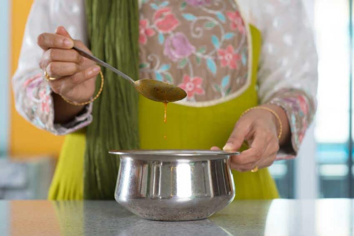 A close-up of a person in a green and white floral garment holding a metal spoon as a golden, orange-tinted liquid drips back into a stainless steel cooking pot.
