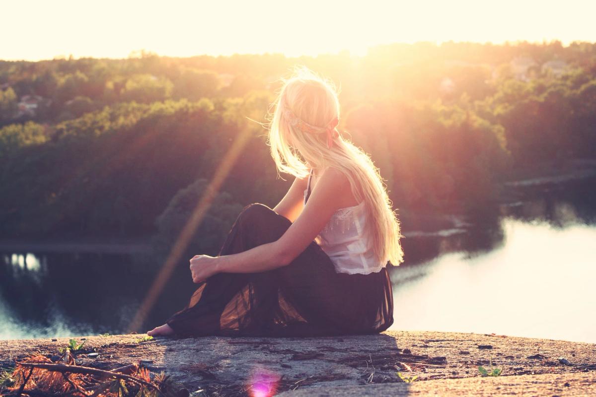 Blonde woman sitting on a cliff at sunset overlooking a calm river and forest landscape, enjoying a peaceful moment of reflection in golden light.