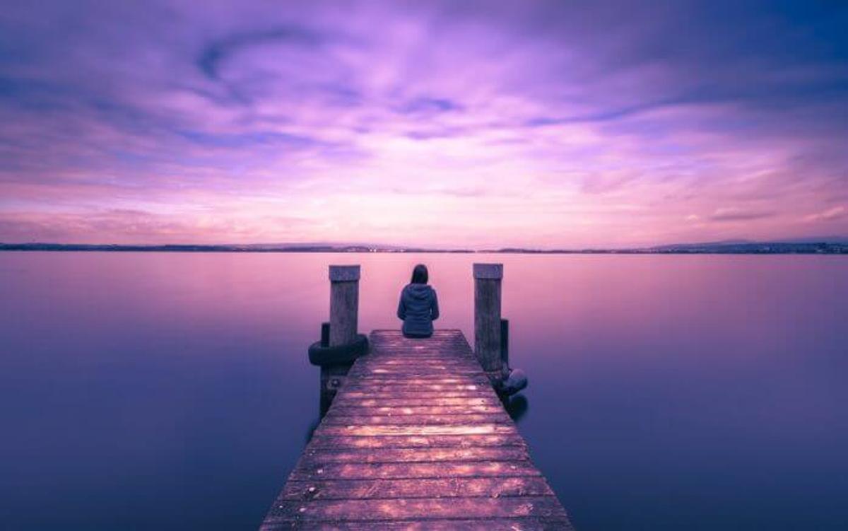 A person sits at the end of a wooden dock facing a calm lake under a purple-pink sunset sky.