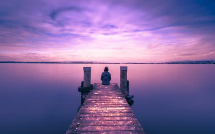 A person sits at the end of a wooden dock facing a calm lake under a purple-pink sunset sky.