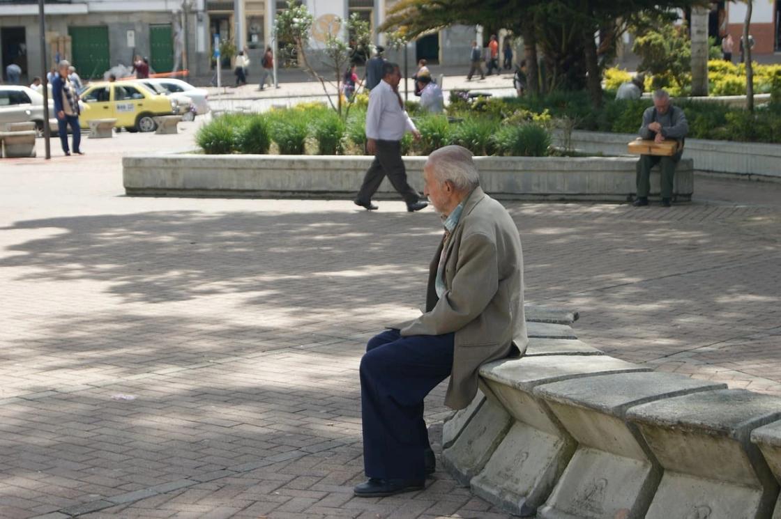 An elderly man in a tan blazer sits alone on a concrete bench in a public plaza.