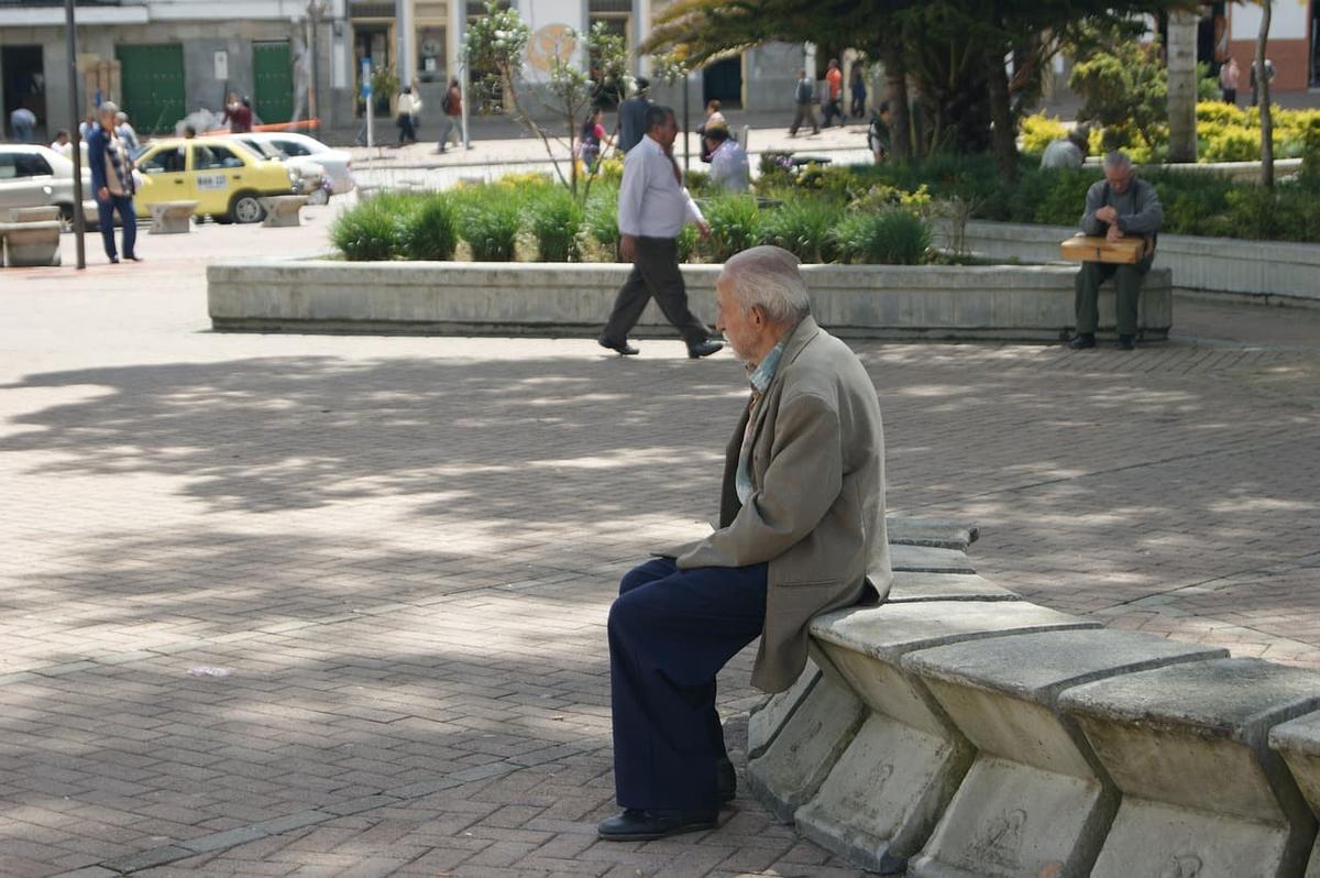 An elderly man in a tan blazer sits alone on a concrete bench in a public plaza.