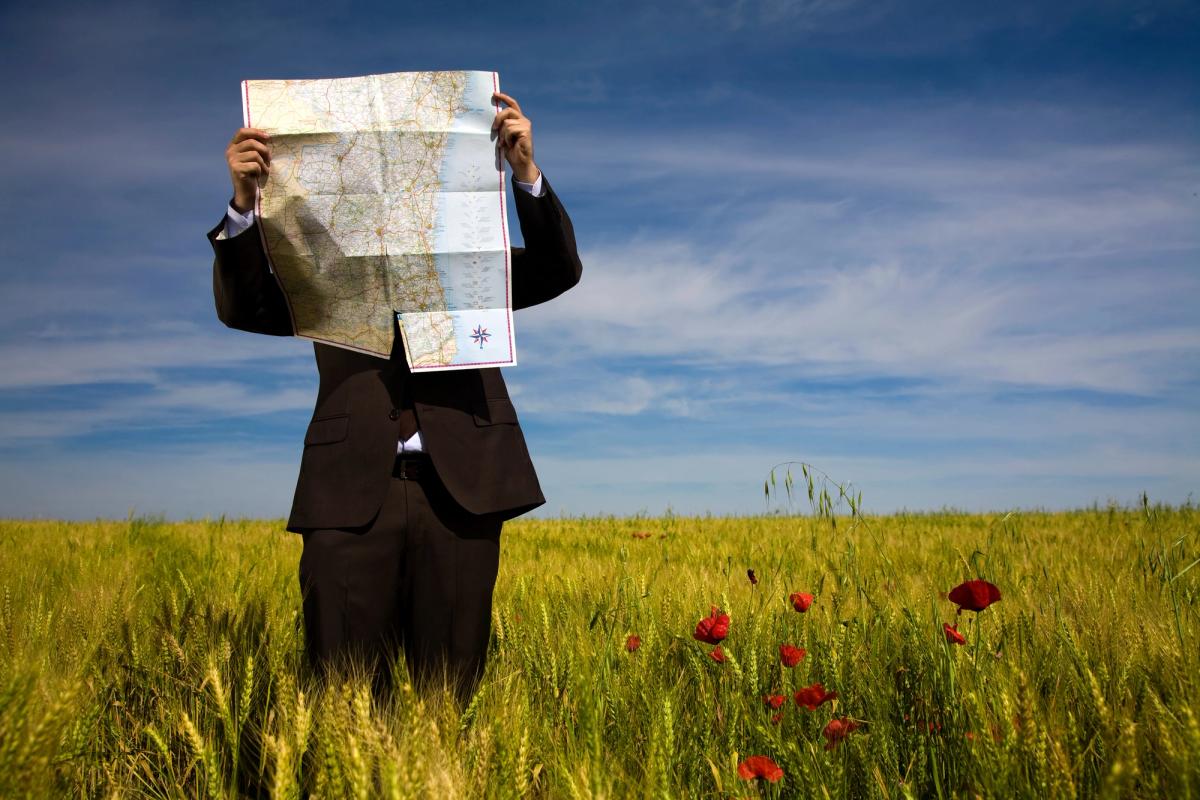A person in a business suit stands in a wide grassy field holding a large paper map up to their face under a blue sky, symbolizing the search for direction and options to find state-funded rehab.