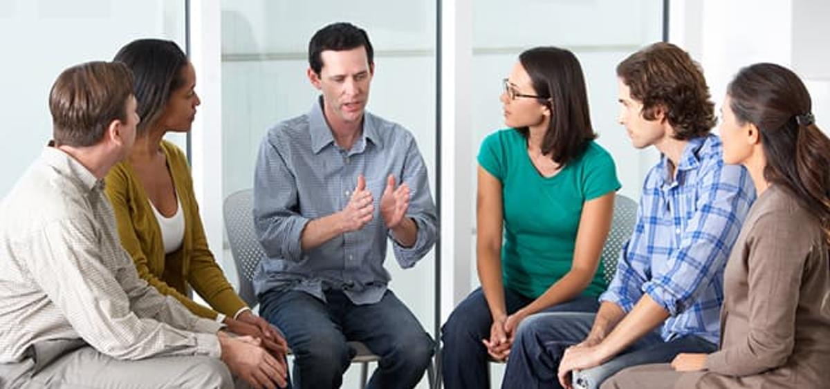 A small group of five adults sits in a circle in a bright room, listening as a man in the center gestures while speaking, suggesting a group therapy or support meeting during 60-day rehab.