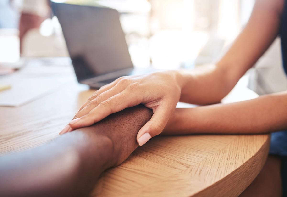 Close-up of one person gently holding another person’s forearm across a wooden table, symbolizing support, compassion, and connection during addiction recovery.