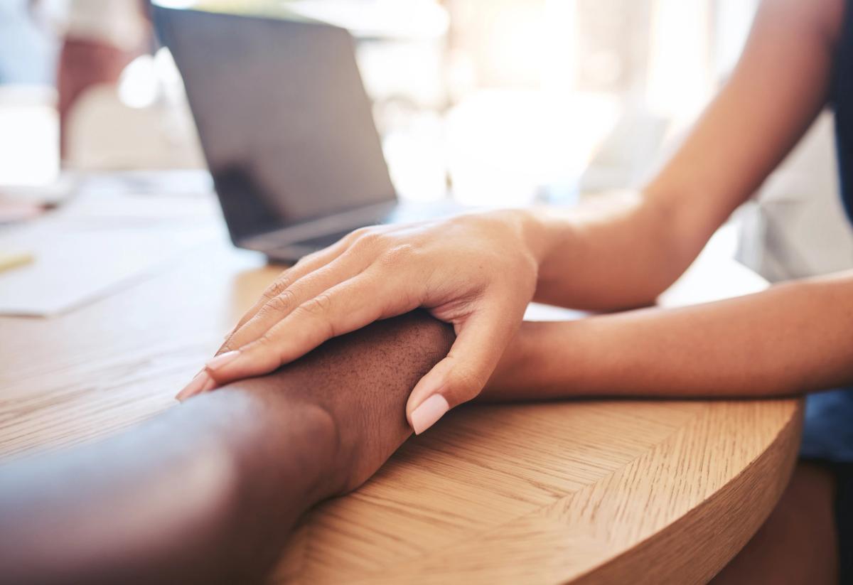 Close-up of one person gently holding another person’s forearm across a wooden table, symbolizing support, compassion, and connection during addiction recovery.