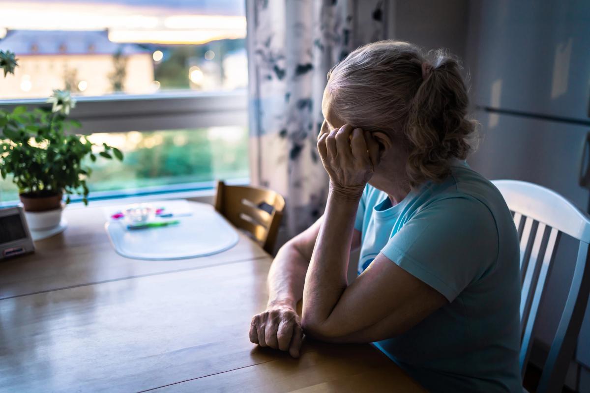 A woman sitting at a kitchen table and looking out a window, resting her head on her hand.