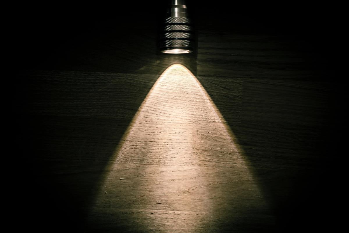 A metal flashlight positioned at the top of the frame, shining a bright triangular beam of warm light downward onto a dark, textured wooden surface.