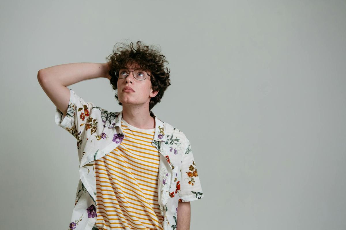 A young person with curly hair and glasses stands against a plain background, one hand resting on the back of their head while looking upward thoughtfully, representing reflection and uncertainty often associated with cross addiction.