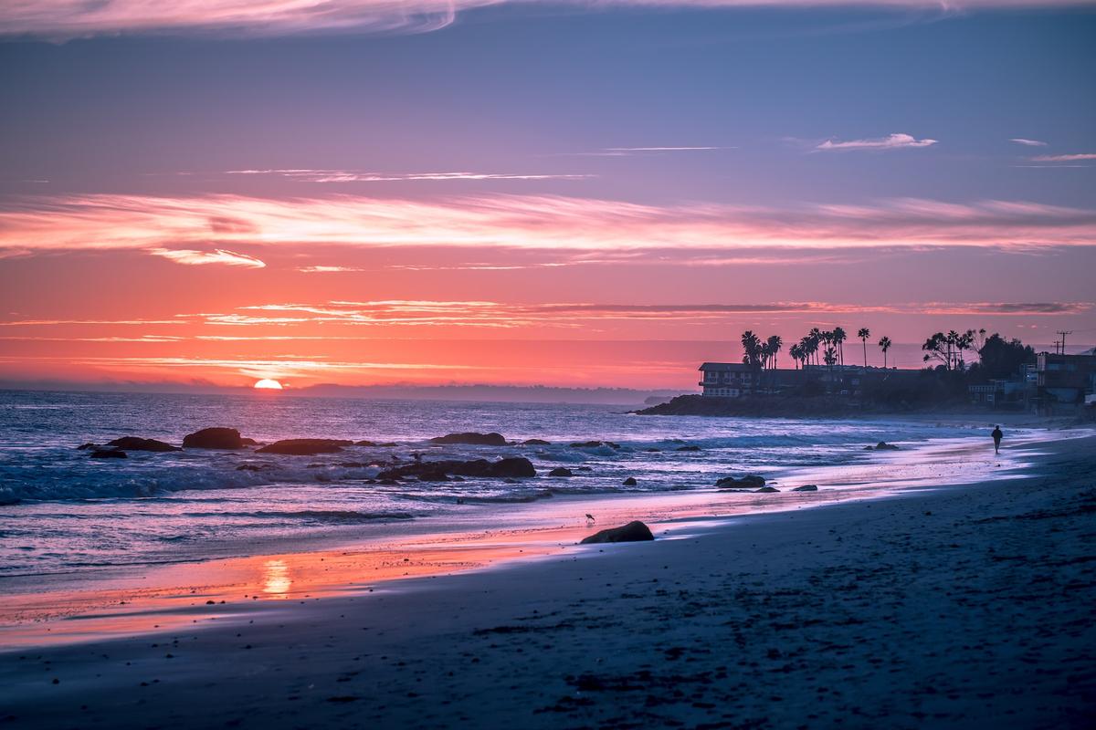 A wide-angle view of a coastal sunset featuring a vibrant sky filled with layers of pink, orange, and purple clouds.