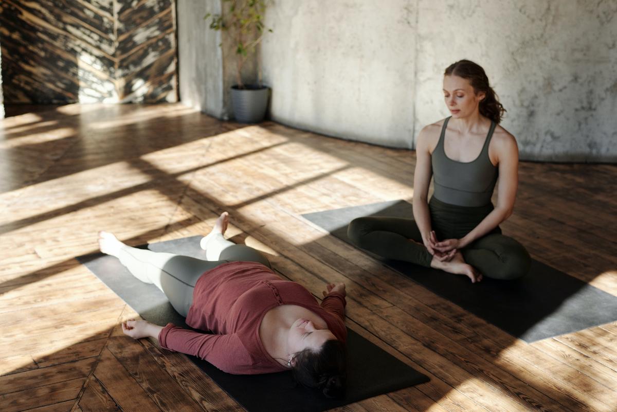 Two women practicing yoga in a sunlit studio; one is lying in Savasana (corpse pose) while the other sits nearby in a cross-legged meditation pose.