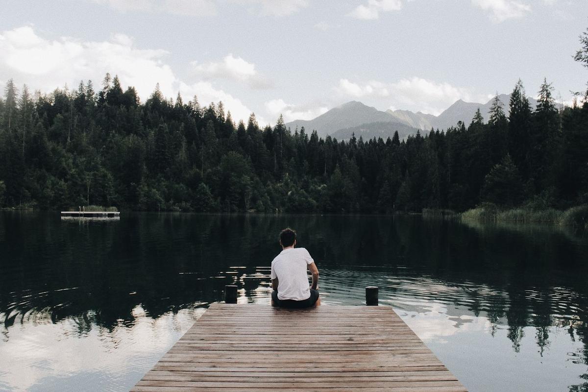 A man in a white t-shirt sits alone at the edge of a wooden dock, looking out over a calm lake surrounded by a dense evergreen forest and distant mountains under a cloudy sky.
