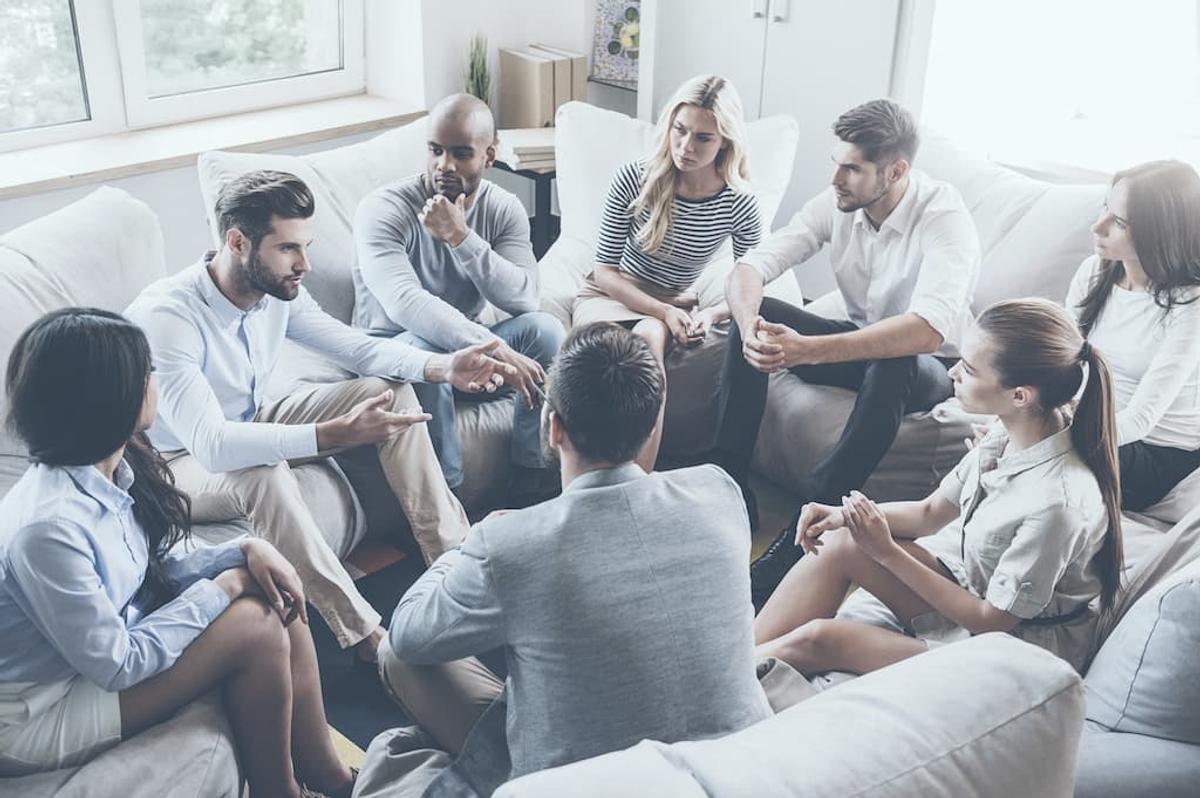 A diverse group of young adults sitting in a circle on beanbag chairs during a collaborative meeting in a bright office.