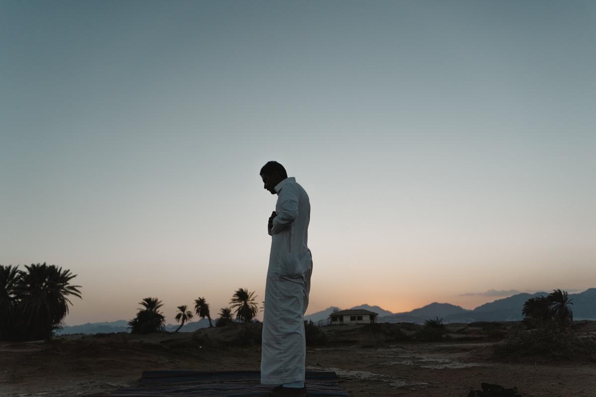 ilhouette of a man praying outdoors at sunset, standing on a prayer mat in a desert landscape with palm trees and mountains in the distance, symbolizing faith, peace, and spiritual reflection.