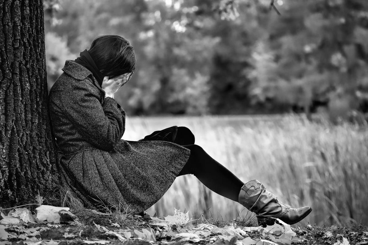 A black and white photo of a person sitting on the ground with their back against a large tree, covering their face with their hands in a gesture of grief or exhaustion.