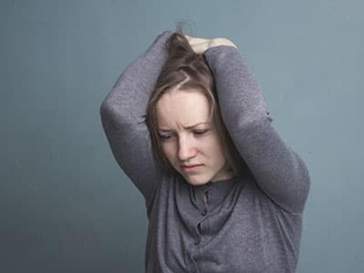 A young woman with a distressed expression holding her head in her hands against a plain grey background.
