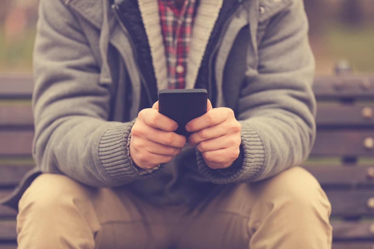 Close-up of a person sitting on a park bench, holding and using a smartphone with both hands