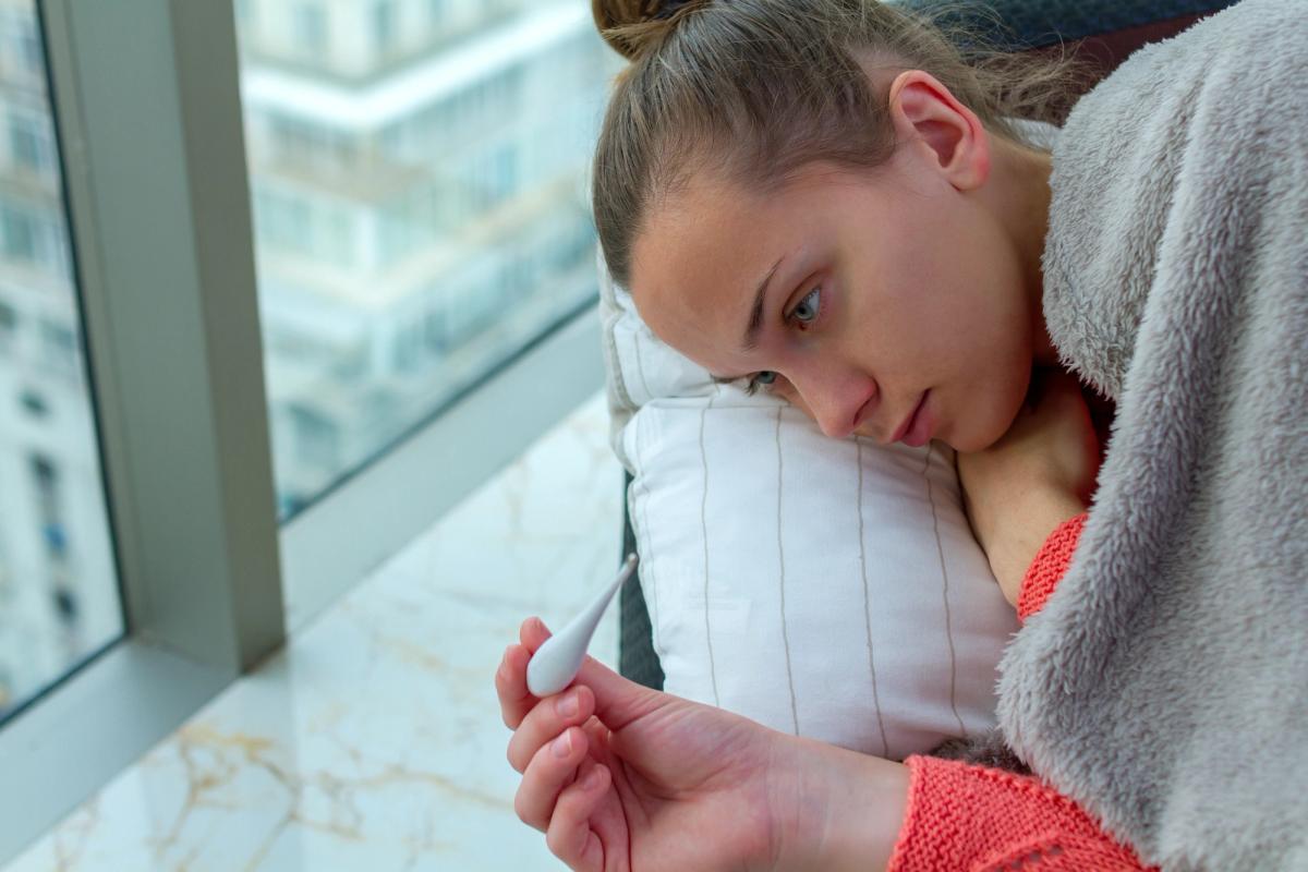 Young woman appearing ill holding thermometer while resting on pillow, illustrating hyperthermia and dangerous health effects of mixing alcohol and cocaine