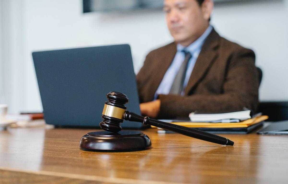 A judge’s gavel rests on a desk in the foreground, with a lawyer in a suit working on a laptop in the background.