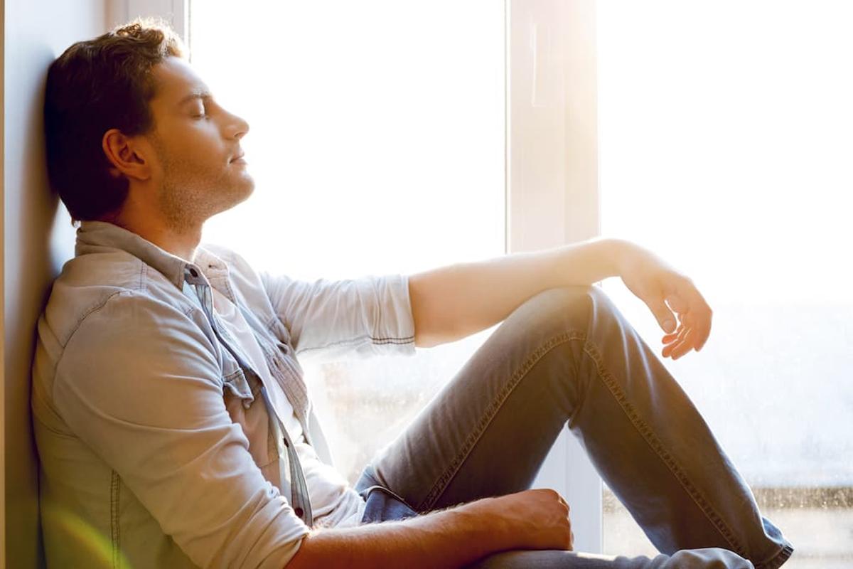 Man sitting on the floor by a bright window with eyes closed, resting and reflecting during recovery or mental health treatment.
