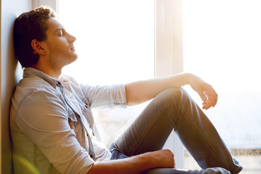 Man sitting on the floor by a bright window with eyes closed, resting and reflecting during recovery or mental health treatment.