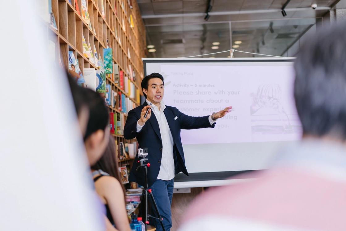 Man in business suit presenting to audience in contemporary office space with bookshelf backdrop, depicting professional success and executive pressure