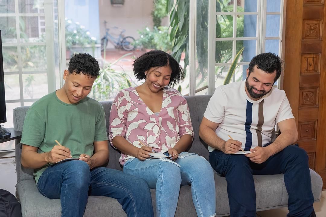 Three people—two men and one woman—sitting side-by-side on a gray sofa, each focused on writing on a small piece of paper with a pencil.