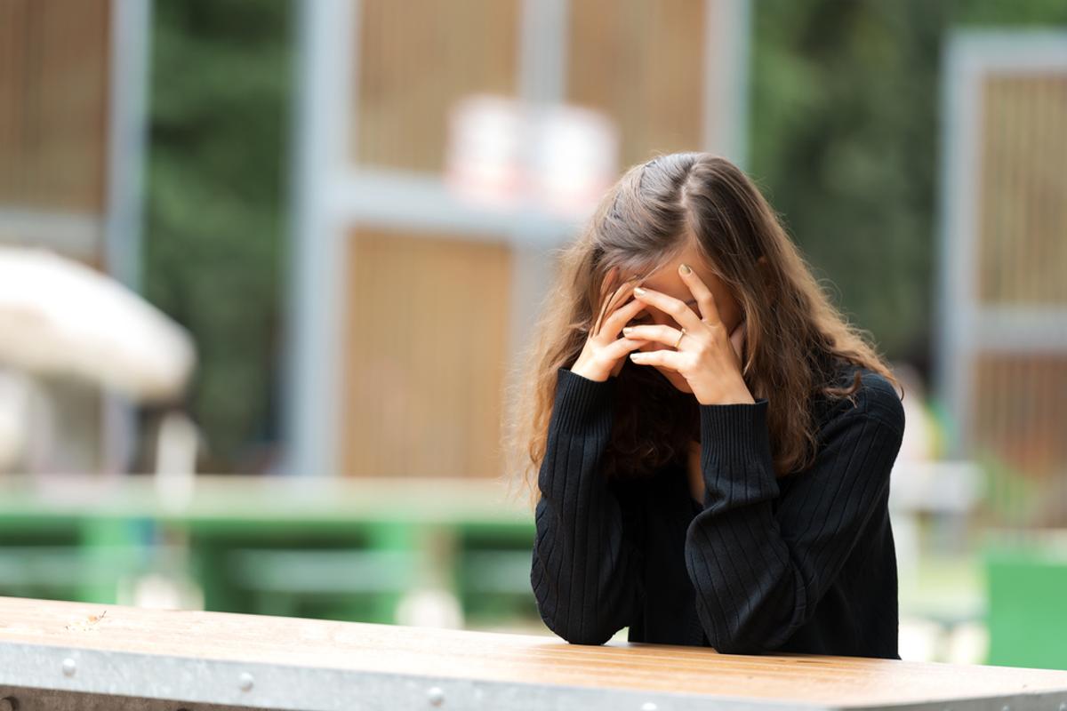 A woman sits outdoors with her head in her hands, appearing distressed or thoughtful.