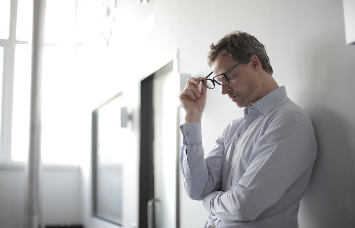 Professional man in business shirt holding glasses with stressed expression in modern office setting, representing executive burnout and need for addiction treatment, bright minimalist workspace with glass doors in background