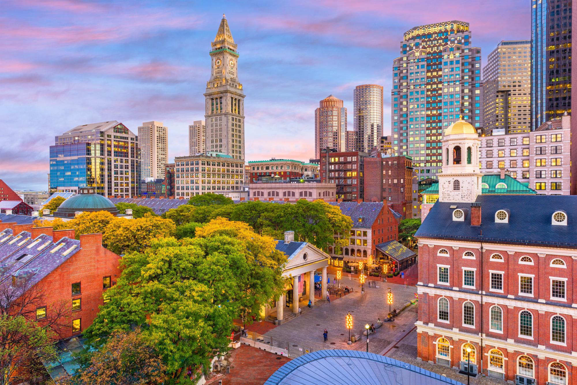 A vibrant city square featuring historic brick buildings with green trees in the foreground, set against a backdrop of modern skyscrapers and the Custom House Tower under a sunset sky