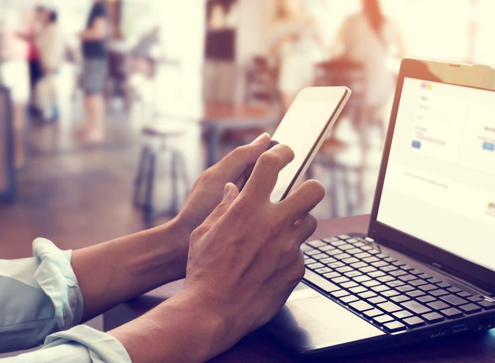 A close-up of a person's hands using a smartphone while sitting at a wooden table next to an open laptop in a bright, blurred cafe setting.