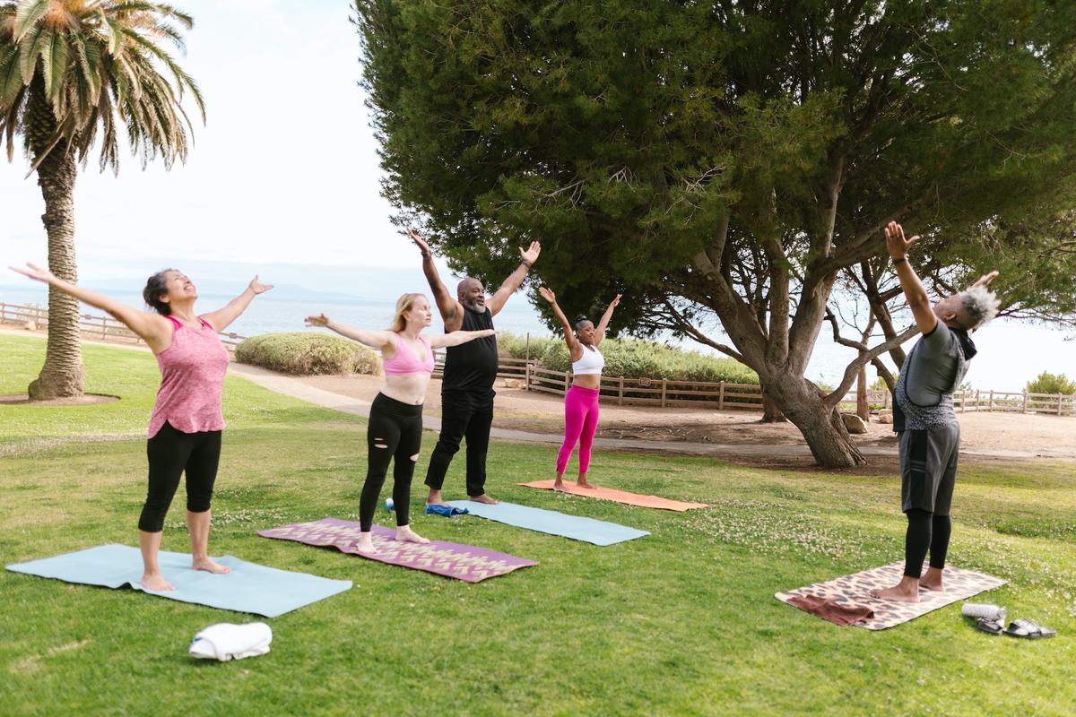 A diverse group of five older adults practicing yoga on a sunny grassy lawn overlooking the ocean.