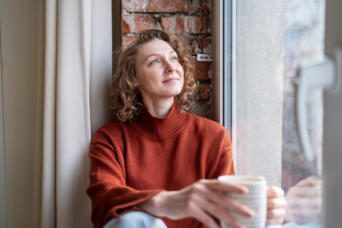 A woman in a rust-colored sweater sits by a window holding a mug, gazing thoughtfully outside with soft natural light on her face.