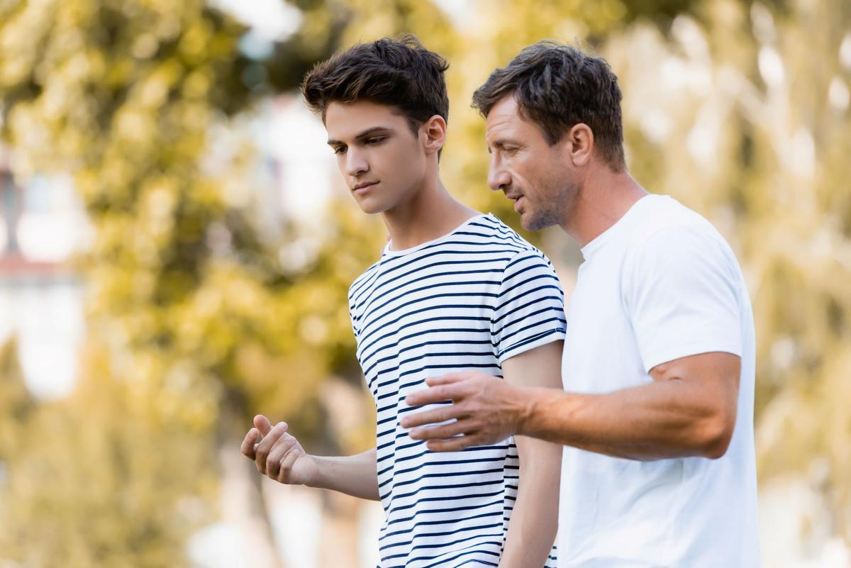 A medium shot of a teenage boy and an older man, likely a father, walking together outdoors in a sun-drenched park.